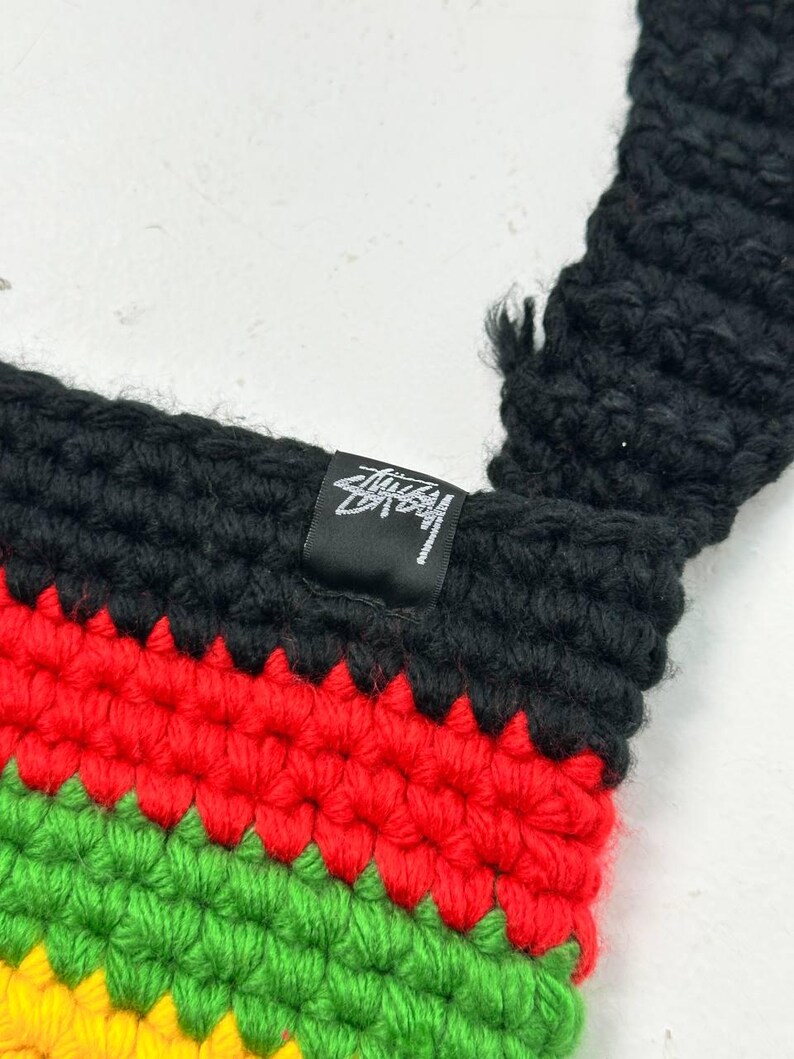 May include: Close-up of a crochet bag with a black strap and a colorful striped pattern. The stripes are red, green, and yellow, with a black top. A small black tag with white text is sewn into the bag.
