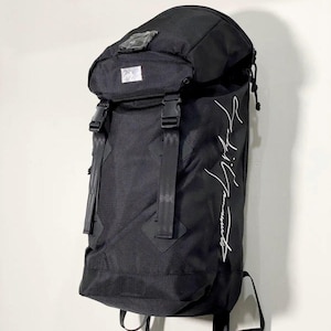 May include: Black backpack with a curved top and adjustable straps. The backpack has a flap closure with buckles and a white logo on the front. The text "Yohji Yamamoto" is written in white on the side.