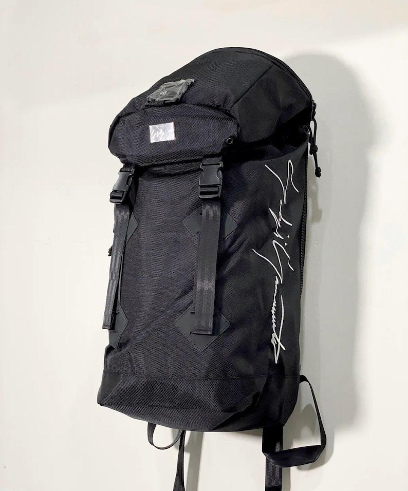May include: Black backpack with a curved top and adjustable straps. The backpack has a flap closure with buckles and a white logo on the front. The text "Yohji Yamamoto" is written in white on the side.