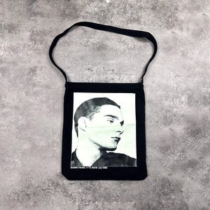 May include: Black tote bag with a black and white portrait of a man. The image is rectangular and centered on the bag. The text "isolated heroes n:10 stevie july 1999" is printed at the bottom of the image.
