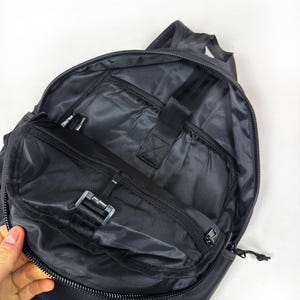 Peut inclure: Un sac &agrave; dos gris fonc&eacute; avec le compartiment principal ouvert, r&eacute;v&eacute;lant une doublure int&eacute;rieure lisse, une poche zipp&eacute;e sur le rabat avant et une sangle &eacute;lastique noire avec une fermeture auto-agrippante pour s&eacute;curiser les objets &agrave; l'int&eacute;rieur.