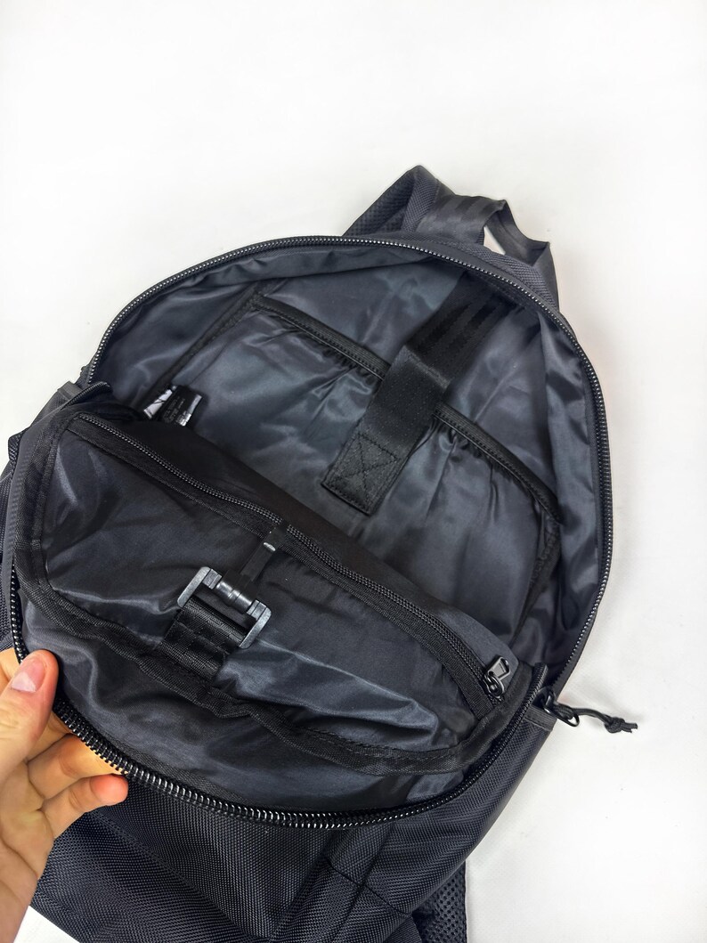 Peut inclure: Un sac &agrave; dos gris fonc&eacute; avec le compartiment principal ouvert, r&eacute;v&eacute;lant une doublure int&eacute;rieure lisse, une poche zipp&eacute;e sur le rabat avant et une sangle &eacute;lastique noire avec une fermeture auto-agrippante pour s&eacute;curiser les objets &agrave; l'int&eacute;rieur.