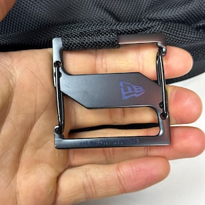 Peut inclure: Une boucle en m&eacute;tal noir avec un cadre rectangulaire et une barre horizontale centrale arborant le logo New Era. Le bord inf&eacute;rieur porte l'inscription grav&eacute;e US PATENT D729,519. La boucle est fix&eacute;e &agrave; une sangle en tissu noir textur&eacute;.