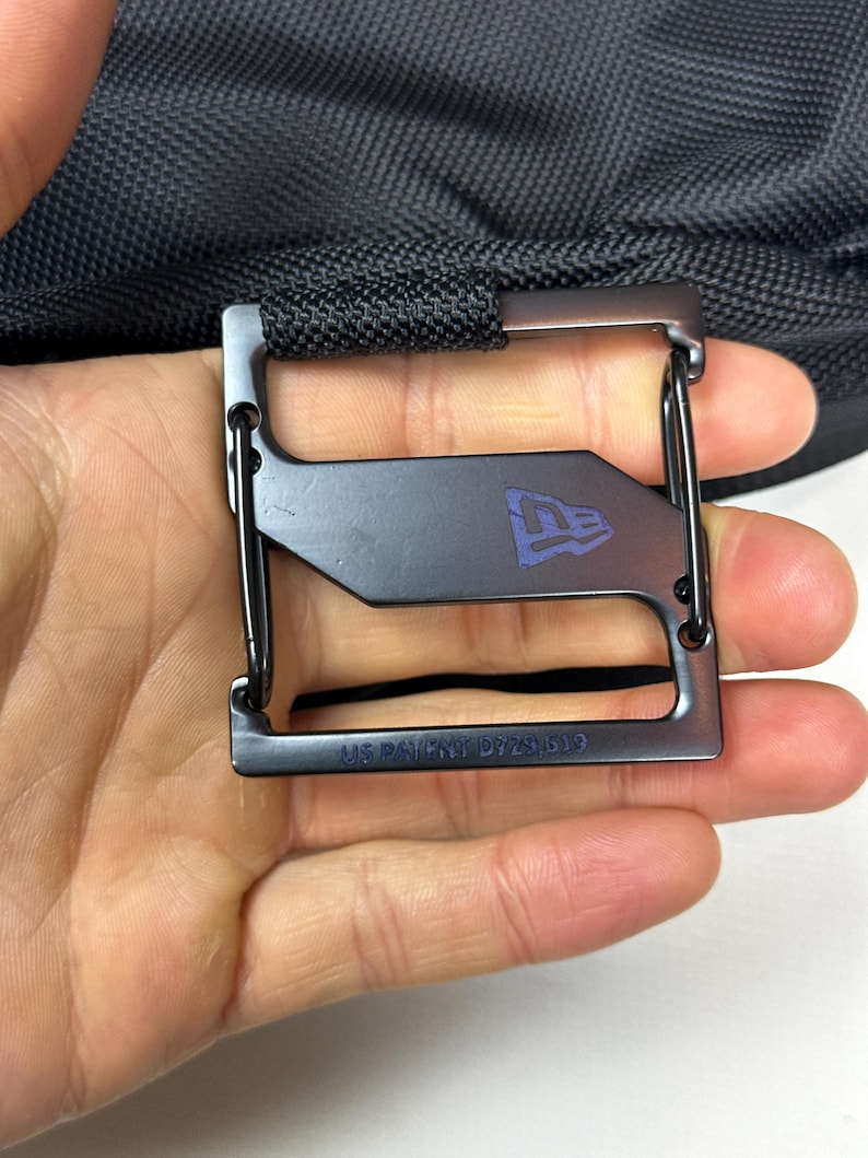 Peut inclure: Une boucle en m&eacute;tal noir avec un cadre rectangulaire et une barre horizontale centrale arborant le logo New Era. Le bord inf&eacute;rieur porte l'inscription grav&eacute;e US PATENT D729,519. La boucle est fix&eacute;e &agrave; une sangle en tissu noir textur&eacute;.