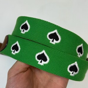 Puede incluir: Un cintur&oacute;n tejido de color verde brillante con un patr&oacute;n repetitivo bordado de s&iacute;mbolos de picas negros delineados con hilo blanco. El cintur&oacute;n tiene una superficie de tela texturizada y un detalle de cuero marr&oacute;n.