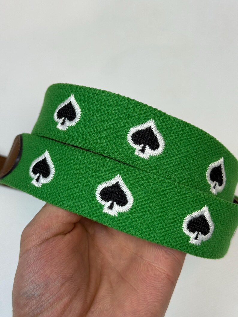 Puede incluir: Un cintur&oacute;n tejido de color verde brillante con un patr&oacute;n repetitivo bordado de s&iacute;mbolos de picas negros delineados con hilo blanco. El cintur&oacute;n tiene una superficie de tela texturizada y un detalle de cuero marr&oacute;n.