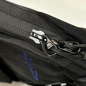 Peut inclure: Un sac noir dot&eacute; d'un ext&eacute;rieur en tissu textur&eacute; avec une fermeture &eacute;clair de style r&eacute;sistant &agrave; l'eau. La tirette est en m&eacute;tal noir, grav&eacute;e du logo YKK, et comprend une boucle en cordon noir pour une prise en main facile. Un motif bleu est visible.