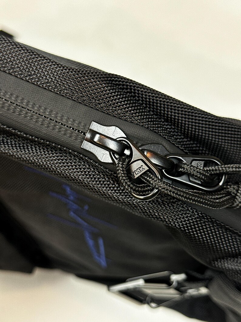 Peut inclure: Un sac noir dot&eacute; d'un ext&eacute;rieur en tissu textur&eacute; avec une fermeture &eacute;clair de style r&eacute;sistant &agrave; l'eau. La tirette est en m&eacute;tal noir, grav&eacute;e du logo YKK, et comprend une boucle en cordon noir pour une prise en main facile. Un motif bleu est visible.