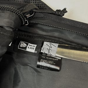 Peut inclure: Un sac en polyester noir avec une &eacute;tiquette de marque New Era et une &eacute;tiquette signature Yohji Yamamoto. L'int&eacute;rieur comprend une &eacute;tiquette d'entretien avec du texte en fran&ccedil;ais et en anglais, pr&eacute;cisant 100% polyester et les instructions.