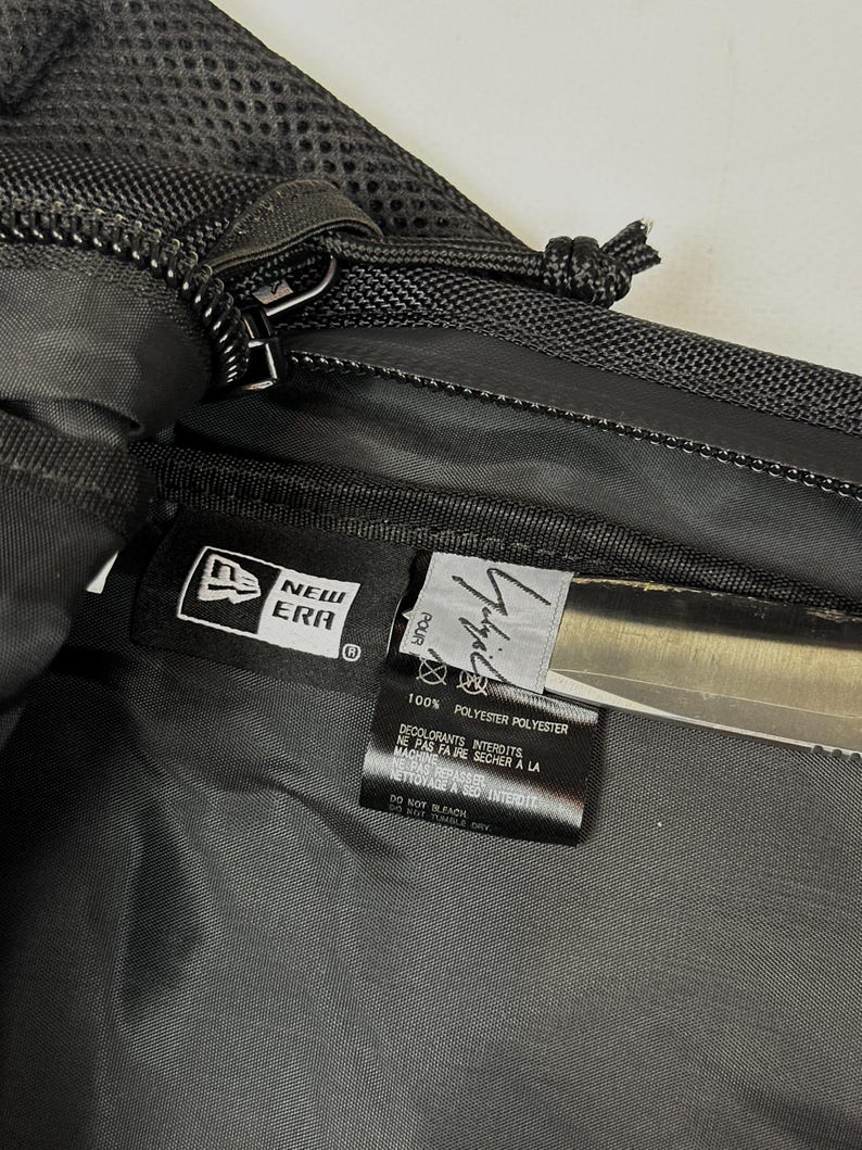 Peut inclure: Un sac en polyester noir avec une &eacute;tiquette de marque New Era et une &eacute;tiquette signature Yohji Yamamoto. L'int&eacute;rieur comprend une &eacute;tiquette d'entretien avec du texte en fran&ccedil;ais et en anglais, pr&eacute;cisant 100% polyester et les instructions.