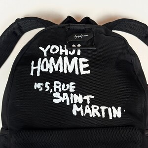Puede incluir: Una mochila negra con texto impreso en blanco que dice YOHJI HOMME 155, RUE SAINT MARTIN. La bolsa incluye un asa superior negra, correas ajustables para los hombros y un detalle de hebilla de metal negro sobre las letras blancas.
