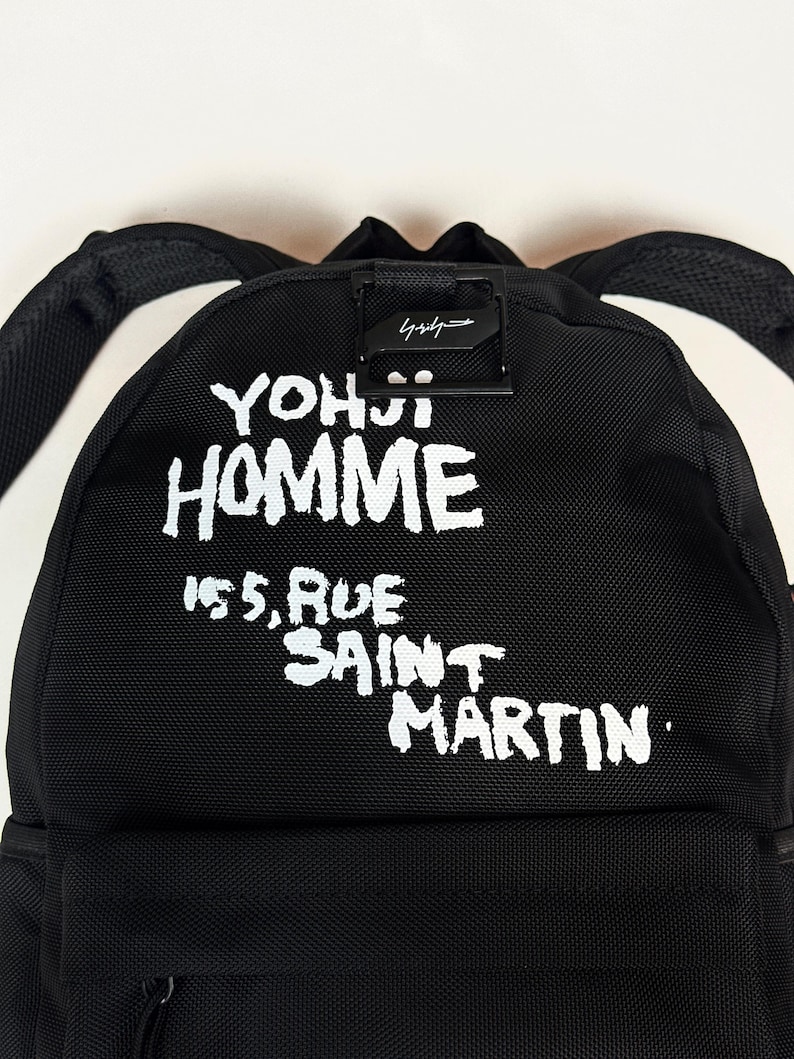 Puede incluir: Una mochila negra con texto impreso en blanco que dice YOHJI HOMME 155, RUE SAINT MARTIN. La bolsa incluye un asa superior negra, correas ajustables para los hombros y un detalle de hebilla de metal negro sobre las letras blancas.