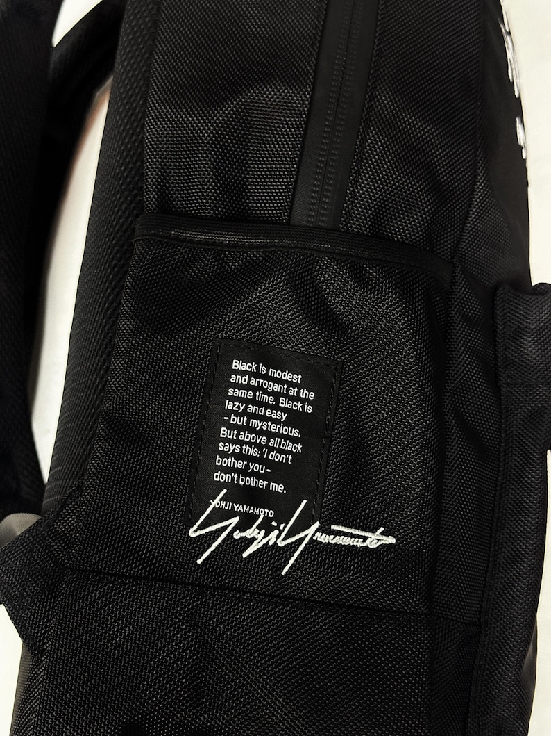 Puede incluir: Una mochila negra que presenta un parche de tela rectangular con texto blanco que dice: Black is modest and arrogant at the same time. Black is lazy and easy - but mysterious. But above all black says this: I don't bother you-don't bother me.