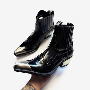 May include: Pair of black leather cowboy boots with silver metal toe caps and heels. The boots have decorative flame-like stitching and a glossy finish. The top of the boots have the text "J'ADIOR".