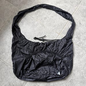 May include: Black shoulder bag made of a lightweight, crinkled material. The bag has a rounded shape with a drawstring closure and a single shoulder strap. A small white triangle logo is visible on the front.