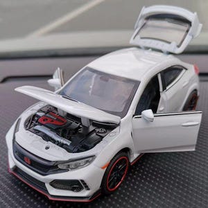 以下が含まれることがあります： 白いモデルカー。ボンネット、ドア、トランクが開いており、エンジンと内装が見える。黒いホイールに赤いアクセント、フロントには赤いホンダのロゴ。詳細なレプリカ。