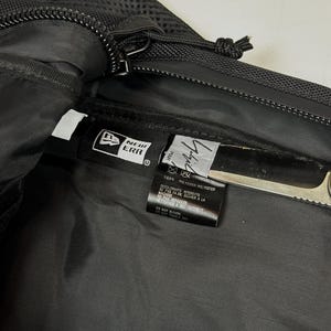 Peut inclure: L'int&eacute;rieur d'un sac en polyester noir avec une &eacute;tiquette de la marque New Era et une &eacute;tiquette secondaire portant le texte Yohji Yamamoto. L'&eacute;tiquette indique 100% polyester et des instructions d'entretien en anglais et en fran&ccedil;ais.