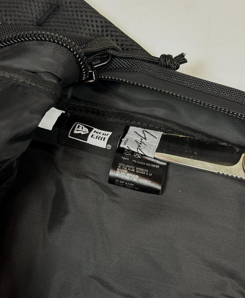 Peut inclure: L'int&eacute;rieur d'un sac en polyester noir avec une &eacute;tiquette de la marque New Era et une &eacute;tiquette secondaire portant le texte Yohji Yamamoto. L'&eacute;tiquette indique 100% polyester et des instructions d'entretien en anglais et en fran&ccedil;ais.
