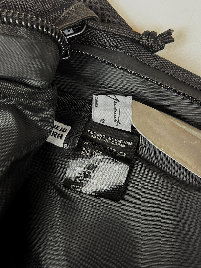 Peut inclure: L'int&eacute;rieur d'un sac en polyester noir avec une &eacute;tiquette de marque New Era et une &eacute;tiquette blanche portant le texte Comme des Gar&ccedil;ons. Une &eacute;tiquette d'entretien noire indique Fabriqu&eacute; au Vietnam, 100% Polyester et Ne pas blanchir.