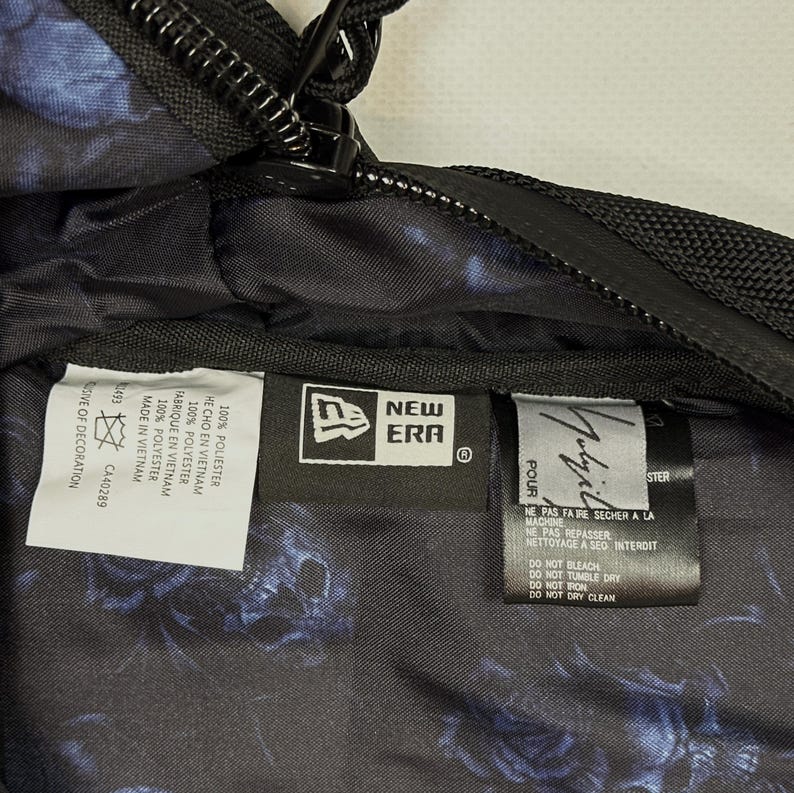 Puede incluir: El interior de una bolsa negra con un patr&oacute;n floral azul y varias etiquetas de tela. Las etiquetas muestran el logotipo de New Era, la marca Yohji Yamamoto e instrucciones de cuidado en ingl&eacute;s, franc&eacute;s y espa&ntilde;ol, incluyendo 100% poli&eacute;ster.