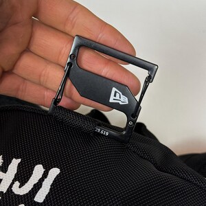 Puede incluir: Un mosquet&oacute;n de metal negro sujeto a una bolsa de tela negra. El mosquet&oacute;n presenta un logotipo de bandera New Era en blanco y el texto impreso 29.619. El clip tiene un marco rectangular con un mecanismo de cierre de resorte.