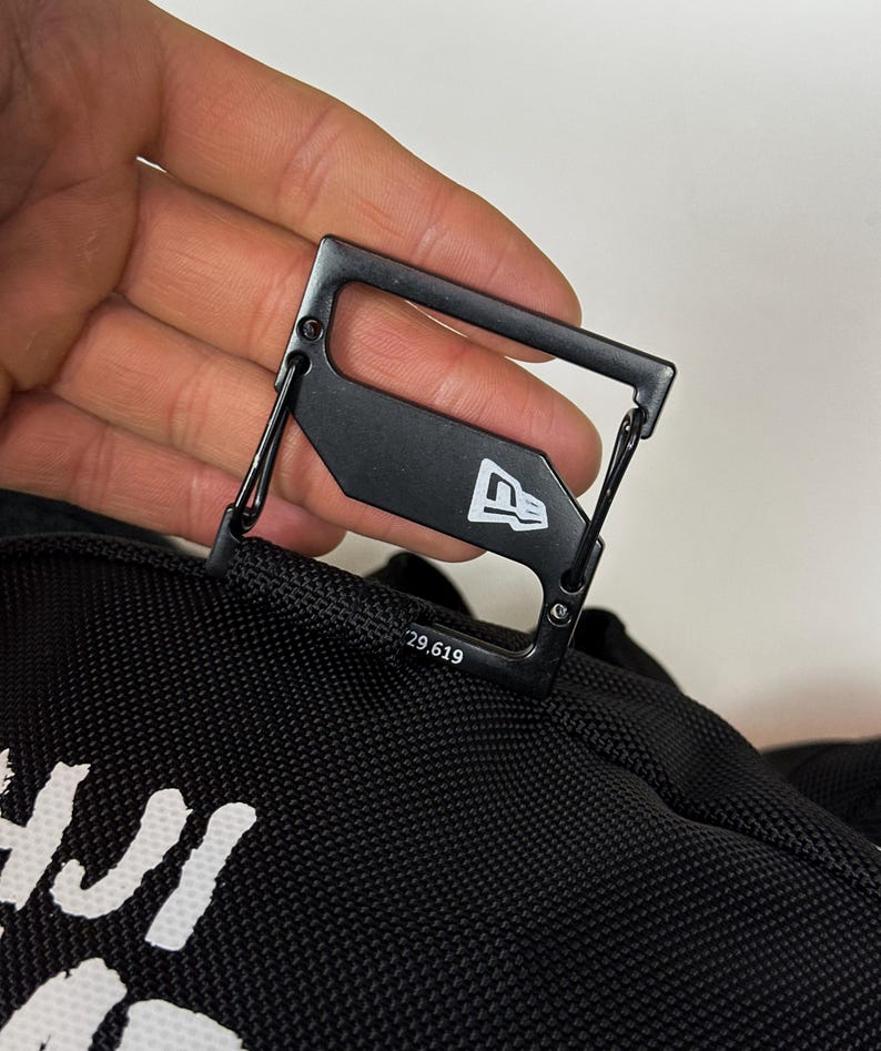 Puede incluir: Un mosquet&oacute;n de metal negro sujeto a una bolsa de tela negra. El mosquet&oacute;n presenta un logotipo de bandera New Era en blanco y el texto impreso 29.619. El clip tiene un marco rectangular con un mecanismo de cierre de resorte.