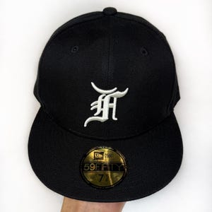 Op de afbeelding: Een zwarte, nauwsluitende honkbalpet met een wit geborduurde gotische letter F op de voorkant. De platte klep is voorzien van een ronde goudkleurige en zwarte sticker met de tekst New Era 59FIFTY 7 1/4, die de specifieke maat aangeeft.