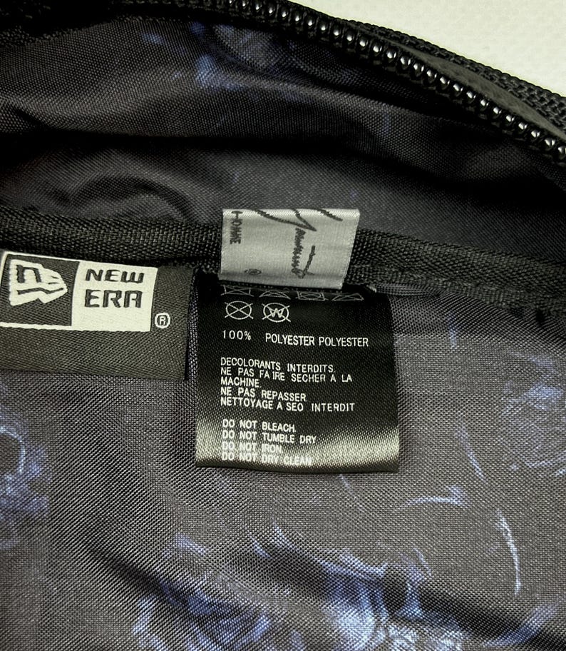 Puede incluir: El interior de una bolsa de poli&eacute;ster negra con una etiqueta de la marca New Era y una etiqueta de cuidado con instrucciones en franc&eacute;s e ingl&eacute;s. La tela tiene un patr&oacute;n abstracto en azul oscuro y negro. La etiqueta indica 100% poli&eacute;ster.