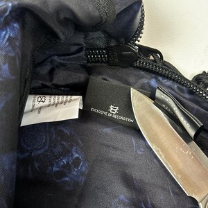 Puede incluir: Un bolso negro con un patr&oacute;n floral azul en el forro interior. Una etiqueta de tela negra en el interior muestra el texto EXCLUSIVE OF DECORATION y un s&iacute;mbolo. Una hoja de cuchillo de metal se coloca sobre la etiqueta, ocultando parcialmente una etiqueta negra secundaria.
