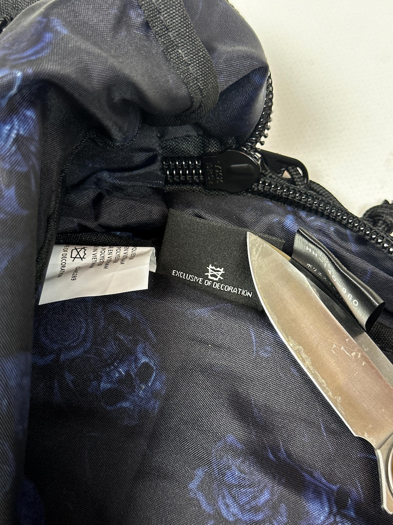 Puede incluir: Un bolso negro con un patr&oacute;n floral azul en el forro interior. Una etiqueta de tela negra en el interior muestra el texto EXCLUSIVE OF DECORATION y un s&iacute;mbolo. Una hoja de cuchillo de metal se coloca sobre la etiqueta, ocultando parcialmente una etiqueta negra secundaria.