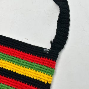 May include: A crochet shoulder bag with a black strap and a body featuring horizontal stripes in red, yellow, green, and black. The bag has a small black tag with white text.