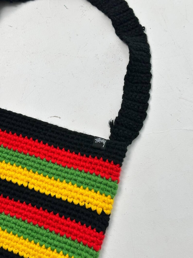May include: A crochet shoulder bag with a black strap and a body featuring horizontal stripes in red, yellow, green, and black. The bag has a small black tag with white text.