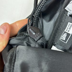 Peut inclure: Un sac en tissu noir dot&eacute; d'une tirette de fermeture &eacute;clair YKK avec une languette en cordon. Une &eacute;tiquette de marque New Era noire et blanche est cousue sur la doublure int&eacute;rieure, avec le mot HOMME visible sur une &eacute;tiquette pr&egrave;s du bord.