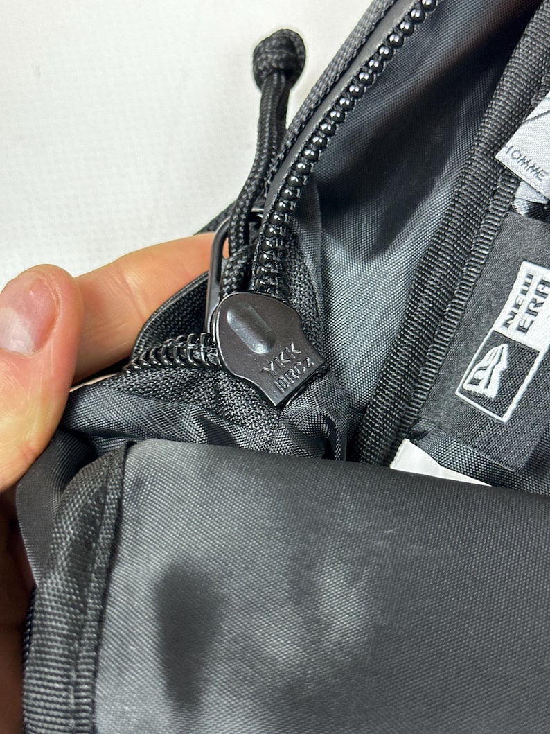 Peut inclure: Un sac en tissu noir dot&eacute; d'une tirette de fermeture &eacute;clair YKK avec une languette en cordon. Une &eacute;tiquette de marque New Era noire et blanche est cousue sur la doublure int&eacute;rieure, avec le mot HOMME visible sur une &eacute;tiquette pr&egrave;s du bord.