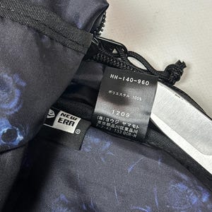 Puede incluir: Un bolso negro con estampado floral azul que presenta una etiqueta con el logo de New Era y una etiqueta interior con el texto HH-140-960, 100% poli&eacute;ster, 1209 y caracteres japoneses que incluyen Yohji Yamamoto y una direcci&oacute;n en Tokio.