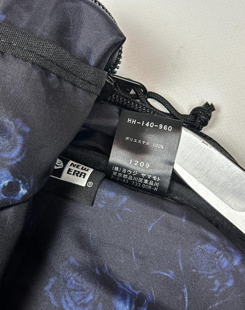 Puede incluir: Un bolso negro con estampado floral azul que presenta una etiqueta con el logo de New Era y una etiqueta interior con el texto HH-140-960, 100% poli&eacute;ster, 1209 y caracteres japoneses que incluyen Yohji Yamamoto y una direcci&oacute;n en Tokio.