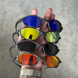 May include: Four pairs of sunglasses with different colored lenses: blue, yellow, black, and orange. The frames are a gunmetal gray color. The sunglasses are held in a hand.