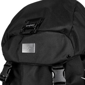 May include: Black backpack with a textured fabric. Features a rectangular black metal buckle and a silver metal plate with a signature logo. The bag has adjustable straps and a top flap.