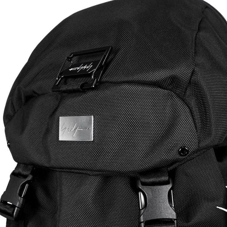 May include: Black backpack with a textured fabric. Features a rectangular black metal buckle and a silver metal plate with a signature logo. The bag has adjustable straps and a top flap.