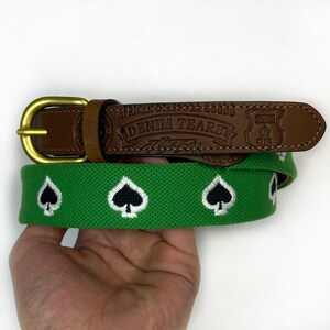 Puede incluir: Un cintur&oacute;n de tela verde con un patr&oacute;n bordado de s&iacute;mbolos de picas en blanco y negro. El cintur&oacute;n incluye un extremo de cuero marr&oacute;n grabado con el texto "AFRICAN DIASPORA GOODS", "DENIM TEARS" y "SHOPS CLUB", asegurado por una hebilla de metal.