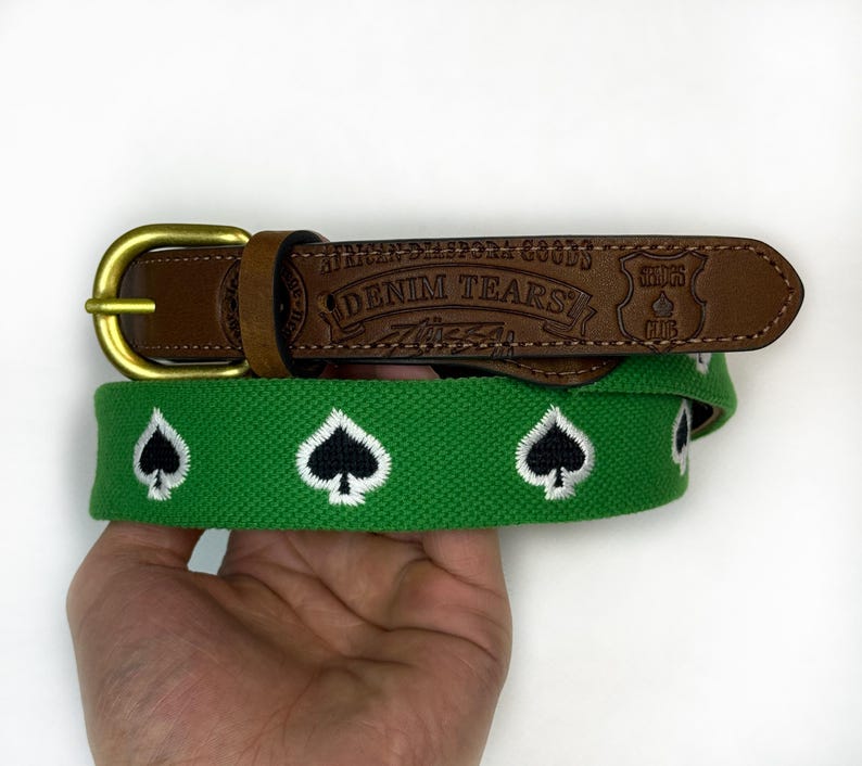 Puede incluir: Un cintur&oacute;n de tela verde con un patr&oacute;n bordado de s&iacute;mbolos de picas en blanco y negro. El cintur&oacute;n incluye un extremo de cuero marr&oacute;n grabado con el texto "AFRICAN DIASPORA GOODS", "DENIM TEARS" y "SHOPS CLUB", asegurado por una hebilla de metal.