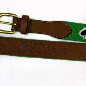 Puede incluir: Un cintur&oacute;n con una correa tejida verde que presenta un s&iacute;mbolo de pica bordado en blanco y negro, unido a un extremo de cuero marr&oacute;n con una hebilla de lat&oacute;n. El cuero tiene grabado el texto African Diaspora Goods, Denim Tears y Spades Club.