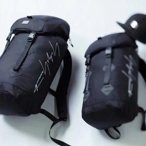 May include: Two black backpacks with the designer's signature in white script. One backpack has a black baseball cap on top. The backpacks have adjustable straps and buckles. The backpacks are made of a durable material.