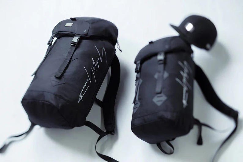 May include: Two black backpacks with the designer's signature in white script. One backpack has a black baseball cap on top. The backpacks have adjustable straps and buckles. The backpacks are made of a durable material.