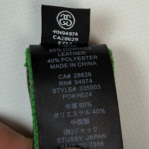 Puede incluir: Una etiqueta de cuidado de prenda de tela negra con texto blanco que presenta el logotipo de Stussy, RN94974, CA28629 y el n&uacute;mero de estilo 335003. La etiqueta indica una composici&oacute;n de 60% cuero vacuno y 40% poli&eacute;ster, fabricado en China.