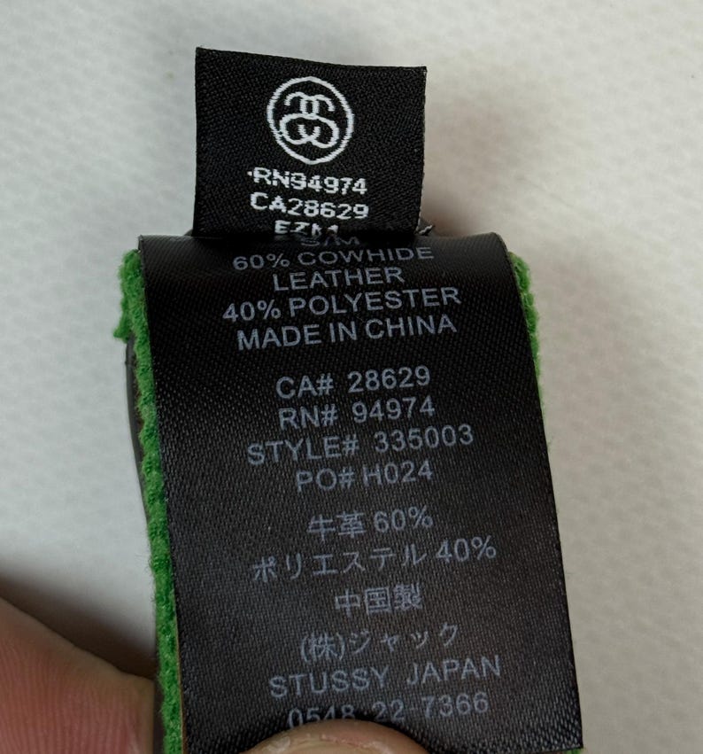 Puede incluir: Una etiqueta de cuidado de prenda de tela negra con texto blanco que presenta el logotipo de Stussy, RN94974, CA28629 y el n&uacute;mero de estilo 335003. La etiqueta indica una composici&oacute;n de 60% cuero vacuno y 40% poli&eacute;ster, fabricado en China.