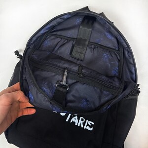Puede incluir: Una mochila negra con el compartimento principal parcialmente abierto que revela un forro interior azul oscuro con un patr&oacute;n floral repetitivo. El interior incluye un bolsillo con cremallera, una correa negra con un gancho y una funda vertical.