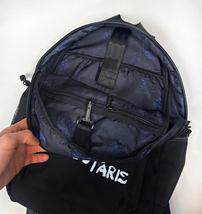 Puede incluir: Una mochila negra con el compartimento principal parcialmente abierto que revela un forro interior azul oscuro con un patr&oacute;n floral repetitivo. El interior incluye un bolsillo con cremallera, una correa negra con un gancho y una funda vertical.