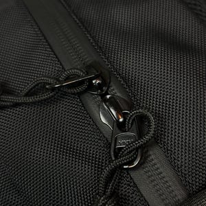 Puede incluir: Una bolsa de tela negra texturizada con cierre de cremallera resistente al agua y dos tiradores de cremallera YKK de metal negro. Cada tirador est&aacute; unido a un lazo de cord&oacute;n negro para un f&aacute;cil agarre, sobre un material sint&eacute;tico resistente.