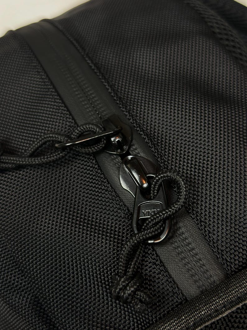 Puede incluir: Una bolsa de tela negra texturizada con cierre de cremallera resistente al agua y dos tiradores de cremallera YKK de metal negro. Cada tirador est&aacute; unido a un lazo de cord&oacute;n negro para un f&aacute;cil agarre, sobre un material sint&eacute;tico resistente.
